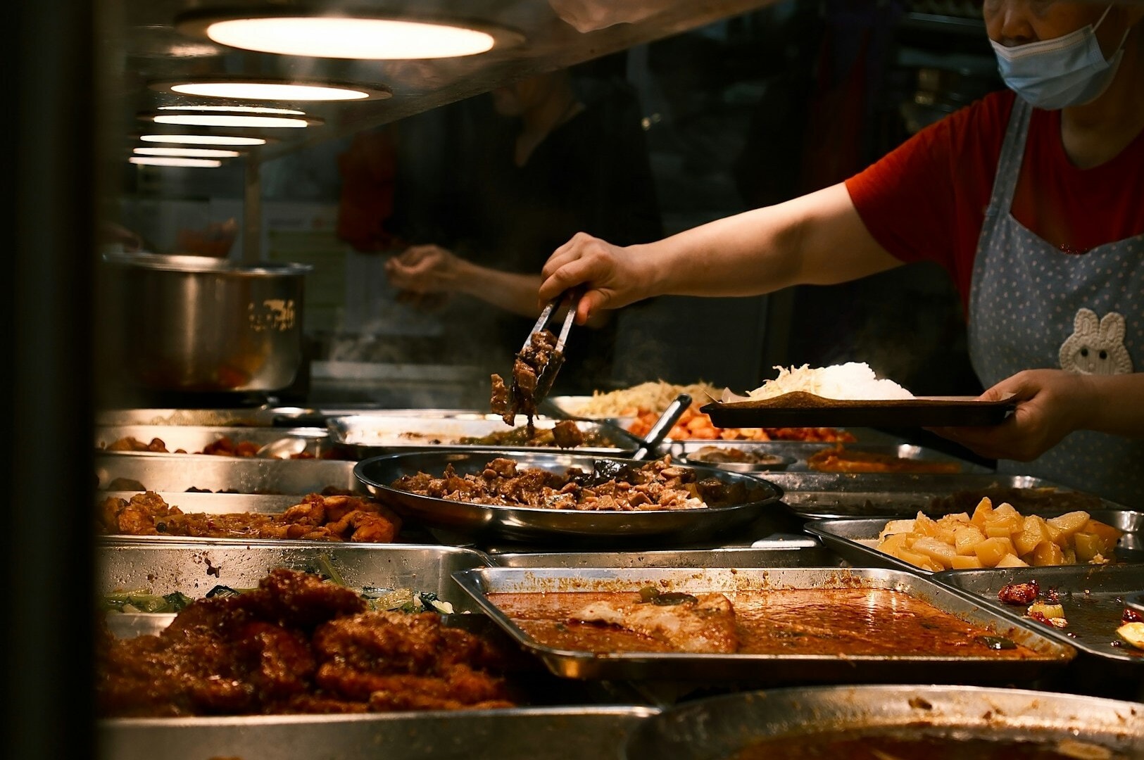 A woman in a mask serves food at a buffet in a Singapore Hawker center, showcasing local culinary delights.