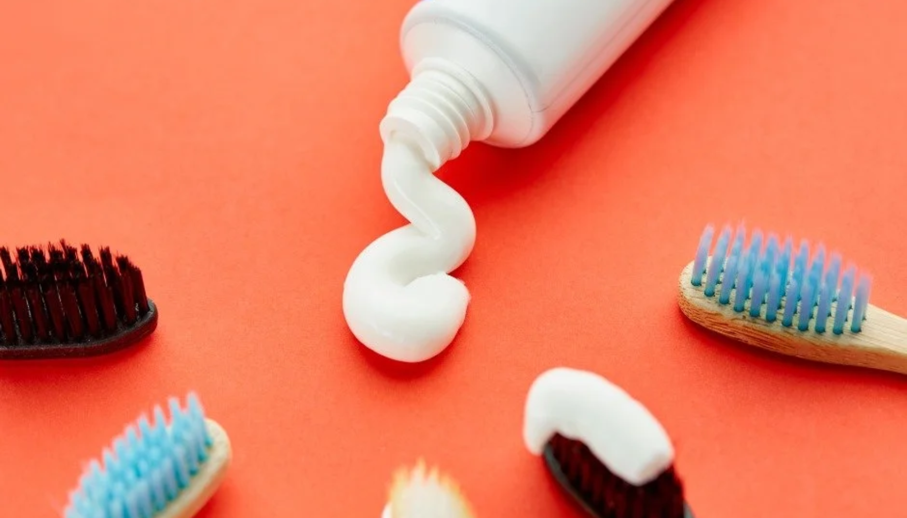 Tannkrem presset ut fra tube omgitt av tannbørster med fargede børstehoder på oransje bakgrunn.