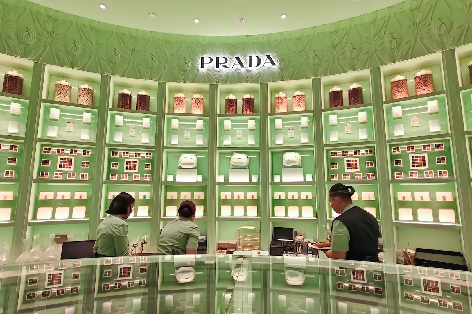 Staff in uniform managing service behind a glass counter at a Prada Cafe surrounded by mint-green floor-to-ceiling merchandise shelving.