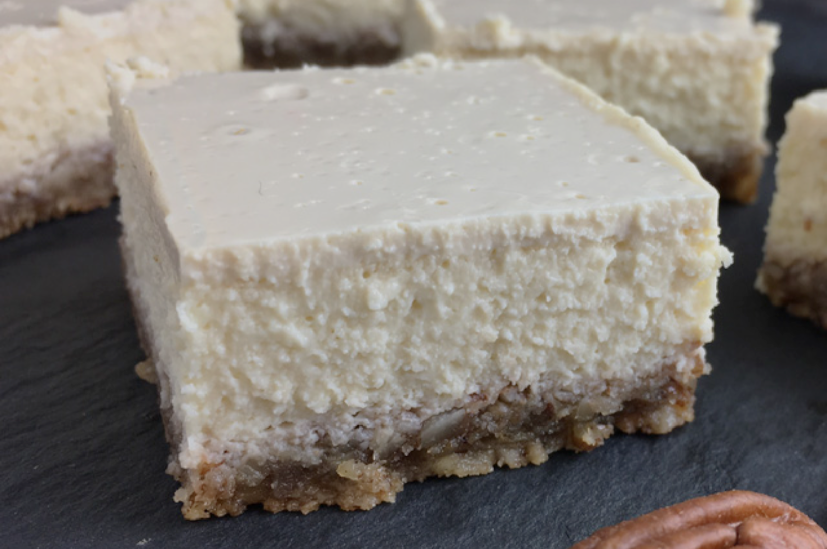 A close-up of a creamy cheesecake bar with a smooth surface and a crumbly, nutty crust on a dark slate background. A pecan is visible nearby.