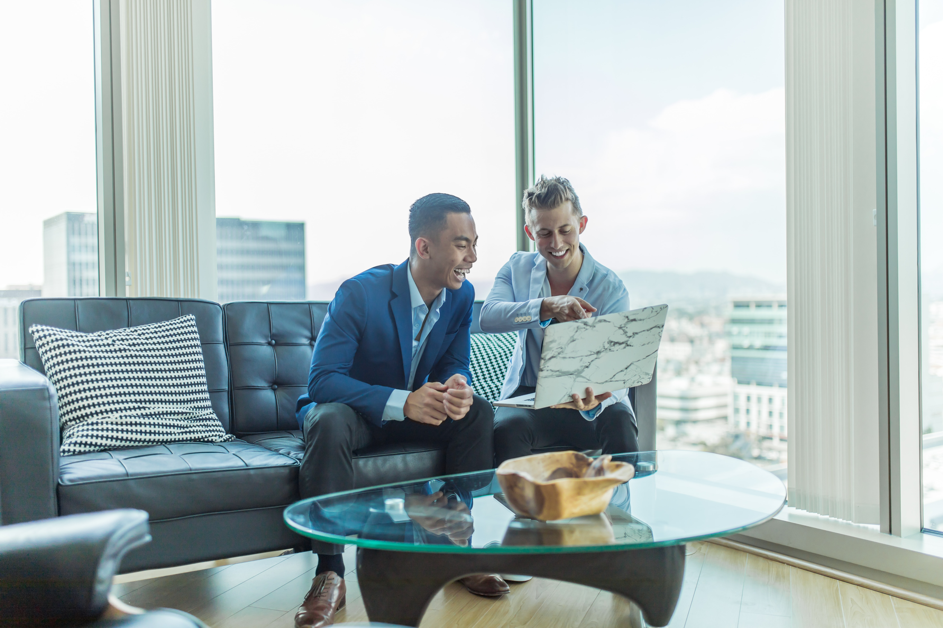 Photo of two men in suit sitting on a sofa while pointing to a laptop.