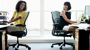 Two female employees sitting in ergonomic office chairs at their desks in a brightly lit office. Both women are smiling and appear engaged in a friendly conversation. The desks are equipped with modern office essentials, including desktop computers, office supplies, and plants. Large windows behind them allow natural light to fill the space, creating a cheerful and inviting work environment. The overall setting emphasizes comfort, ergonomics, and a positive workplace atmosphere.