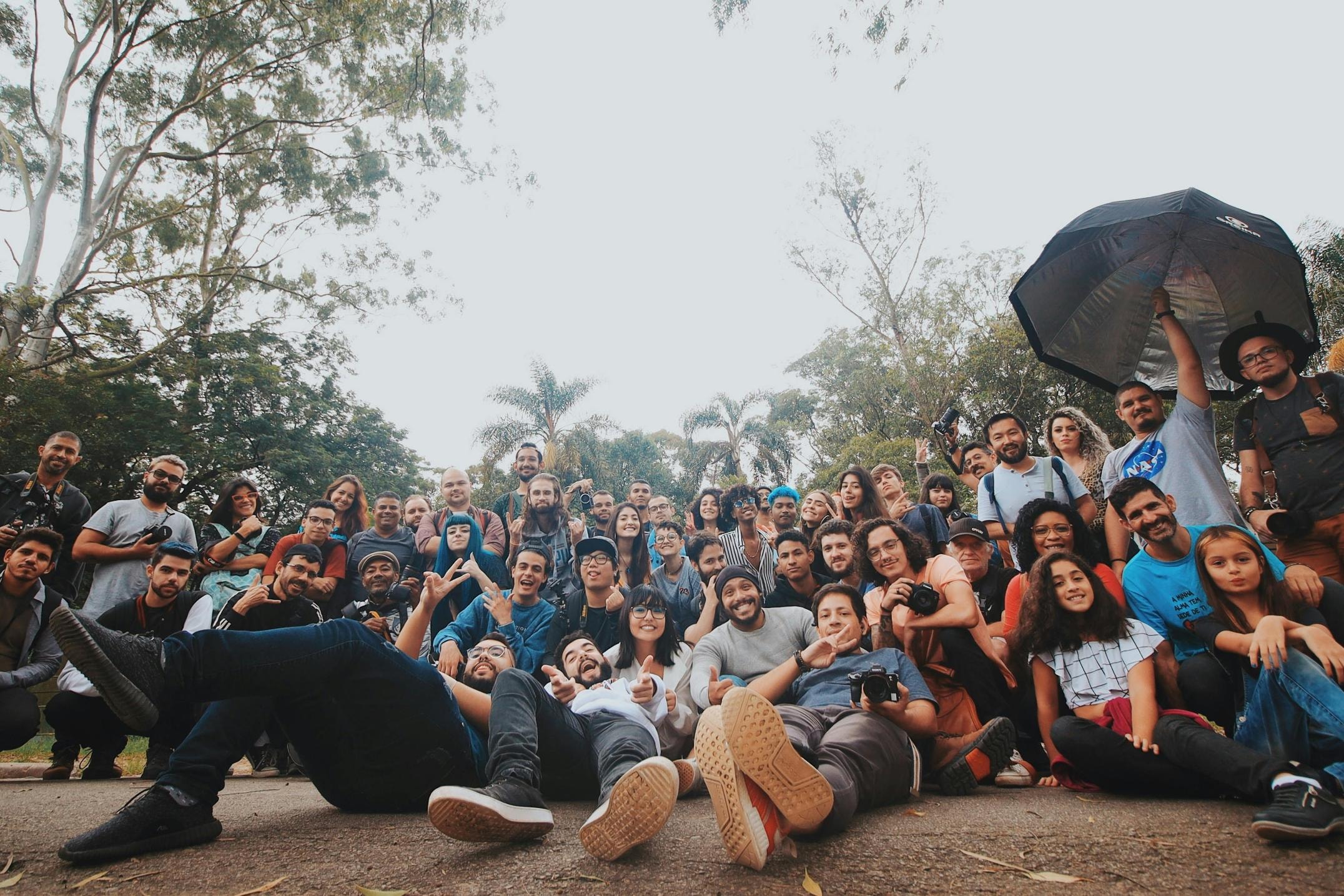 This image features a large, cheerful group of people gathered outdoors, many of whom are holding cameras, suggesting a photography club or meetup. The photo is taken from a distinct low angle, framing the smiling subjects against a backdrop of trees and sky while highlighting a relaxed and communal atmosphere.