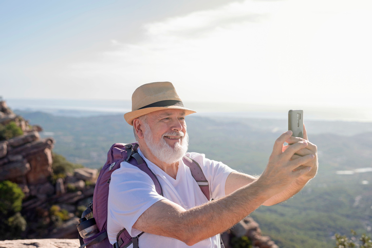 Mature man in a straw hat and backpack snapping a photo with his cell from a mountaintop.