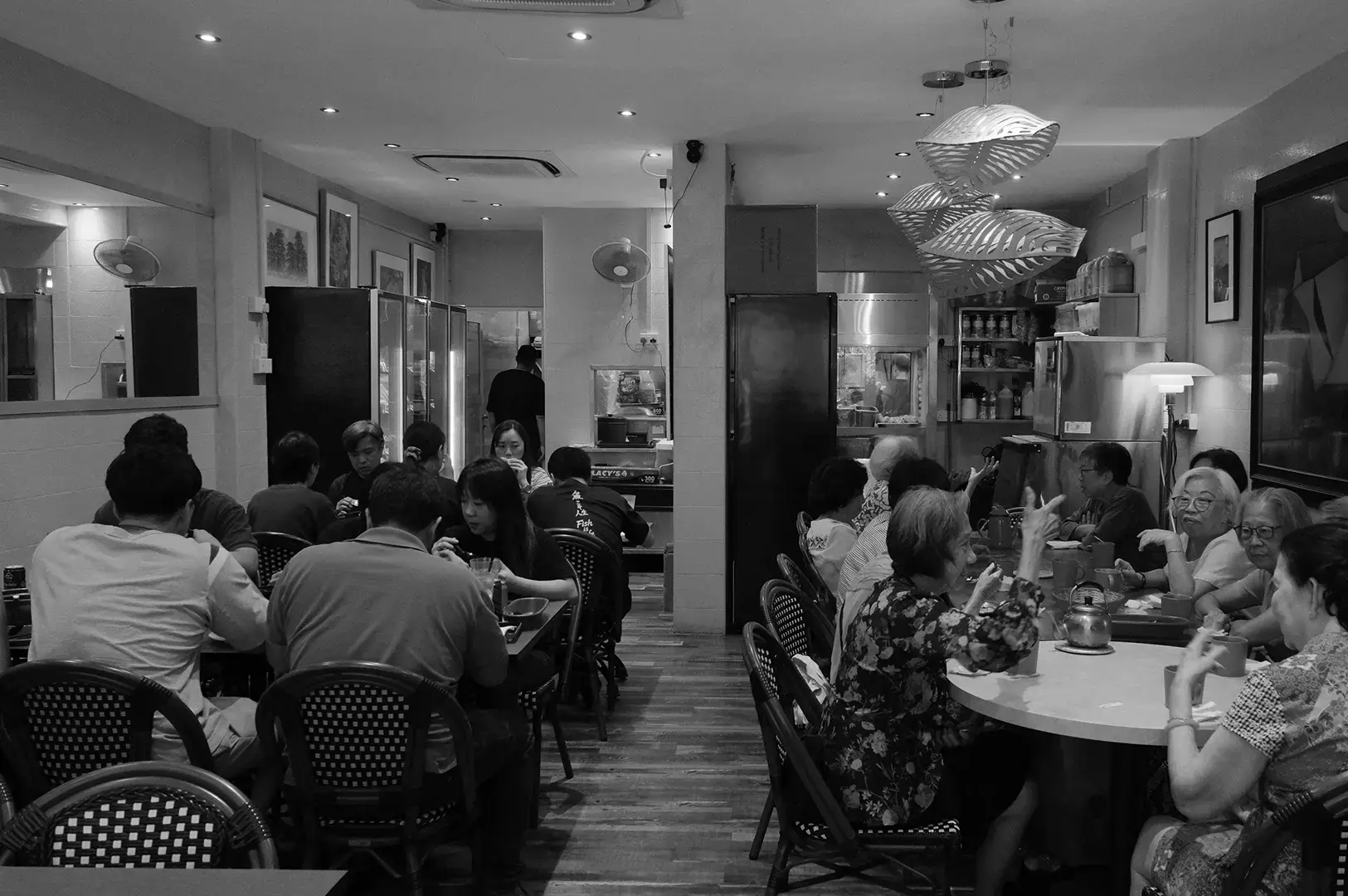 A busy traditional restaurant interior with customers dining at tables, ceiling lights, woven chairs, and an open kitchen area.