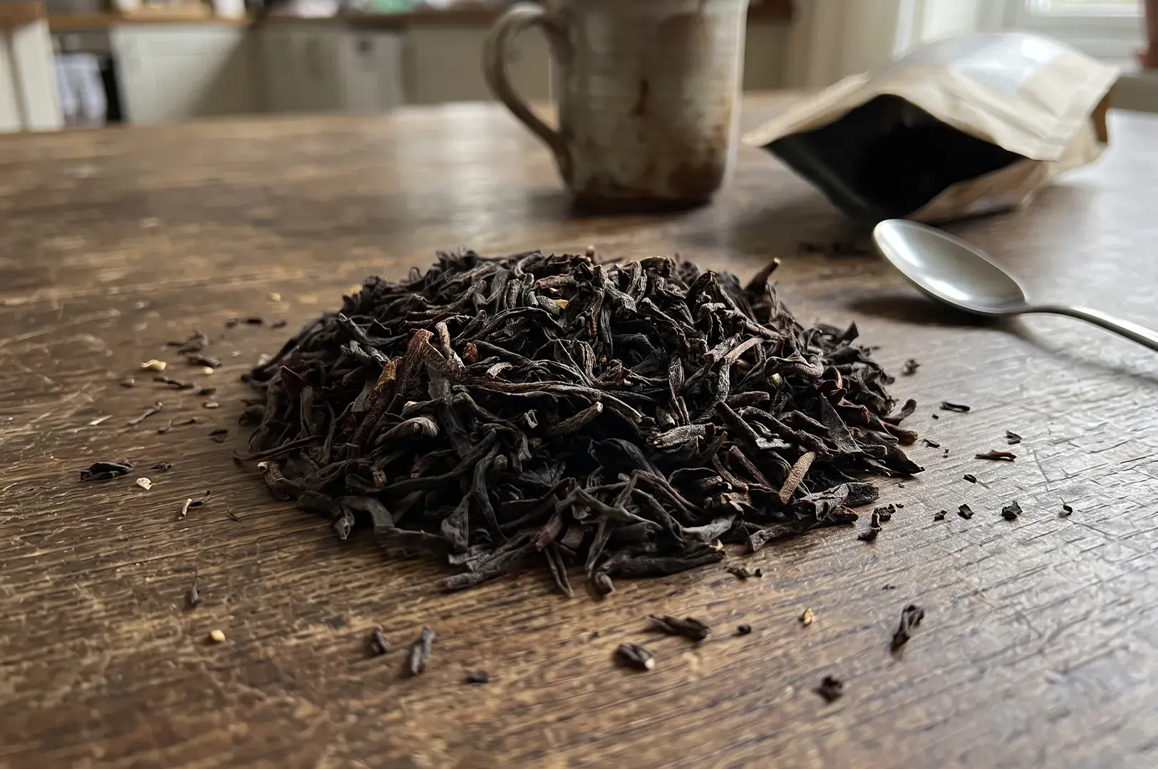 Mound of dark loose-leaf tea placed on a wooden tabletop next to a metal spoon, ceramic mug, and opened tea pouch.