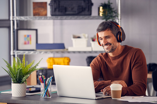Cheerful man in a brown sweater working on his computer with headphones on. 