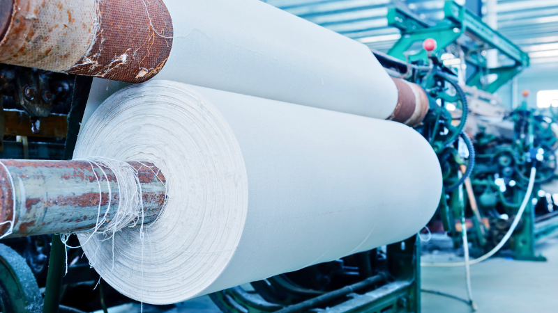 A large roll of white polyester fabric is on a loom machine with other textile machinery in the background.