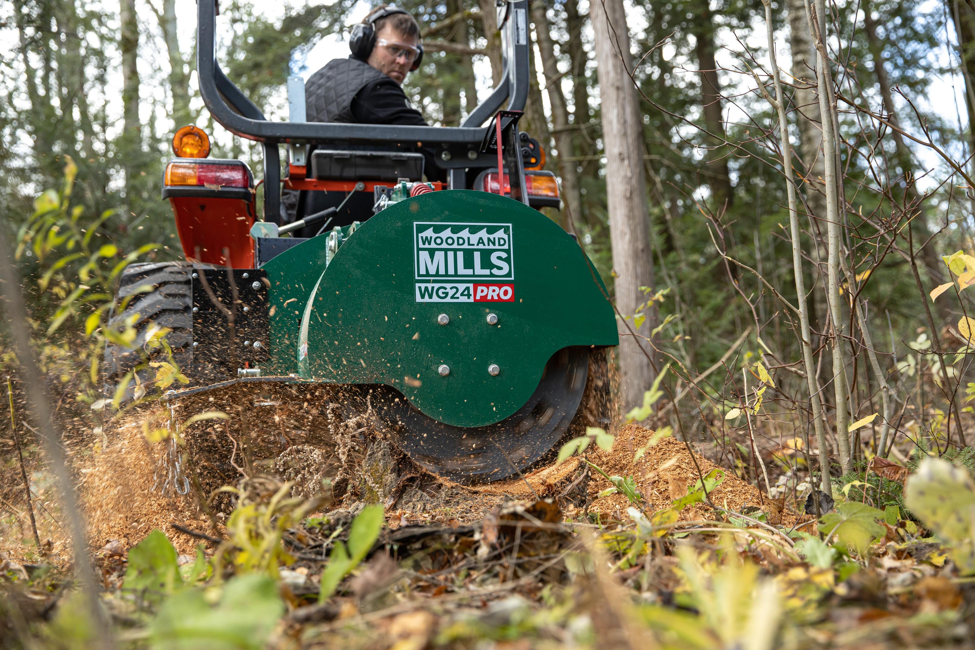 A Woodland Mills WG24 PRO Stump Grinder being drawn through a stump with a man looking over his shoulder on a tractor. 