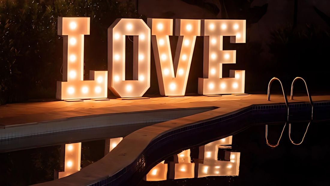  Large illuminated marquee letters spelling “LOVE” placed poolside at night, reflecting in the water and glowing warmly against a dark outdoor backdrop.