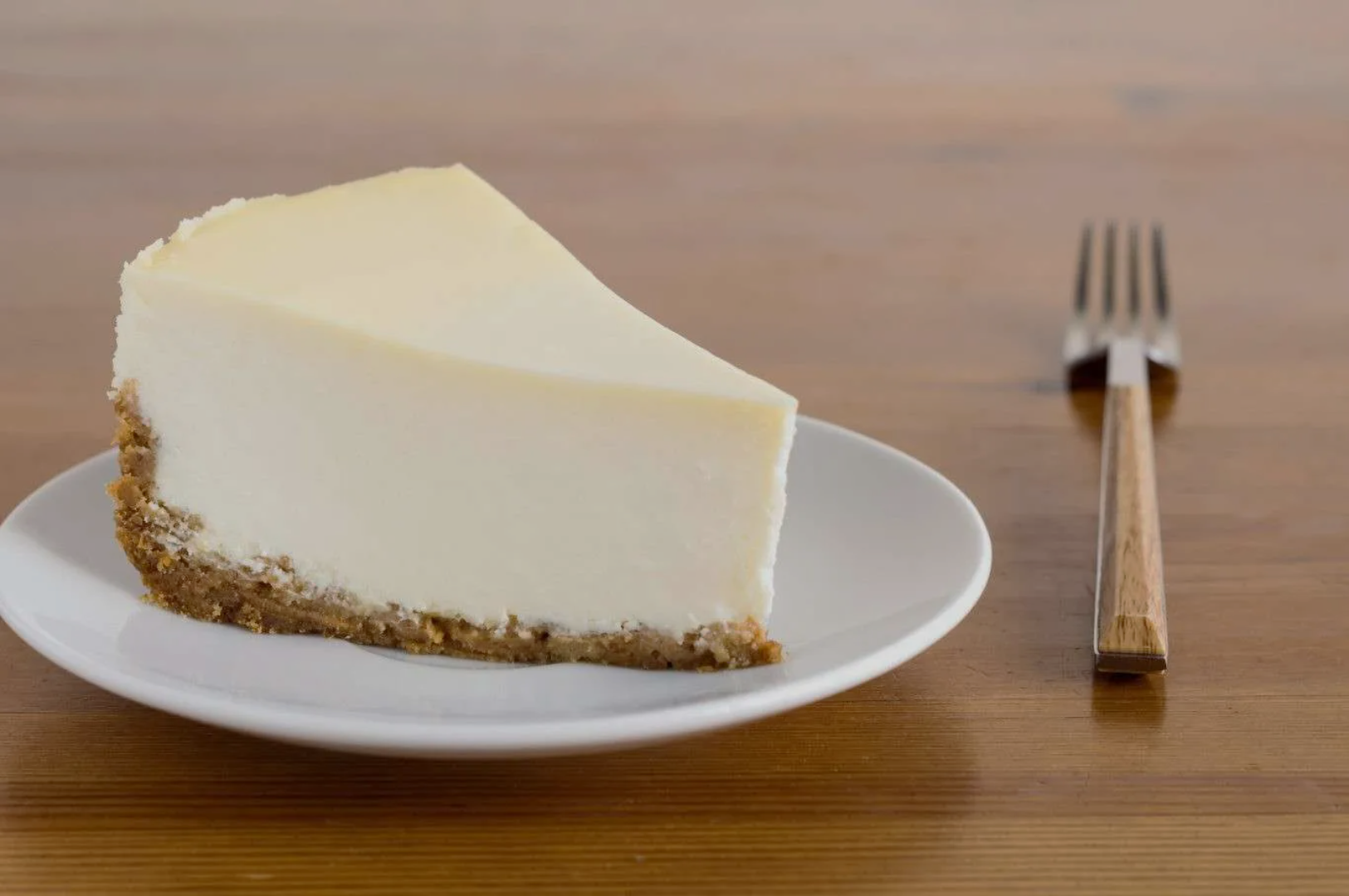 A slice of creamy cheesecake with a graham cracker crust sits on a white plate. A wooden-handled fork lies beside it on a wooden table.