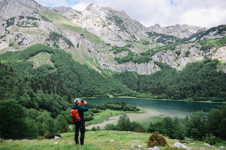 Man with an orange backpack snapping a photo of the mountains and stream. 