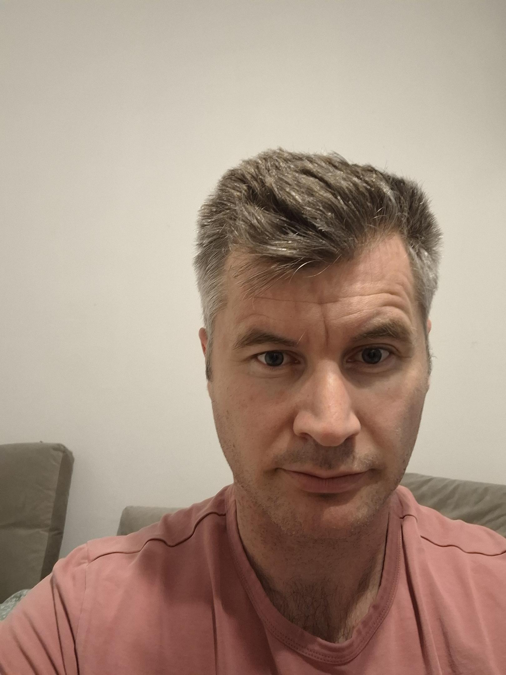 A man in his late 30s to early 40s looking directly into the camera with a neutral, composed expression, wearing a pink t-shirt, with salt-and-pepper hair styled upward, seated in front of a light-colored wall — the close framing and steady gaze suggest a profile or identity photo.