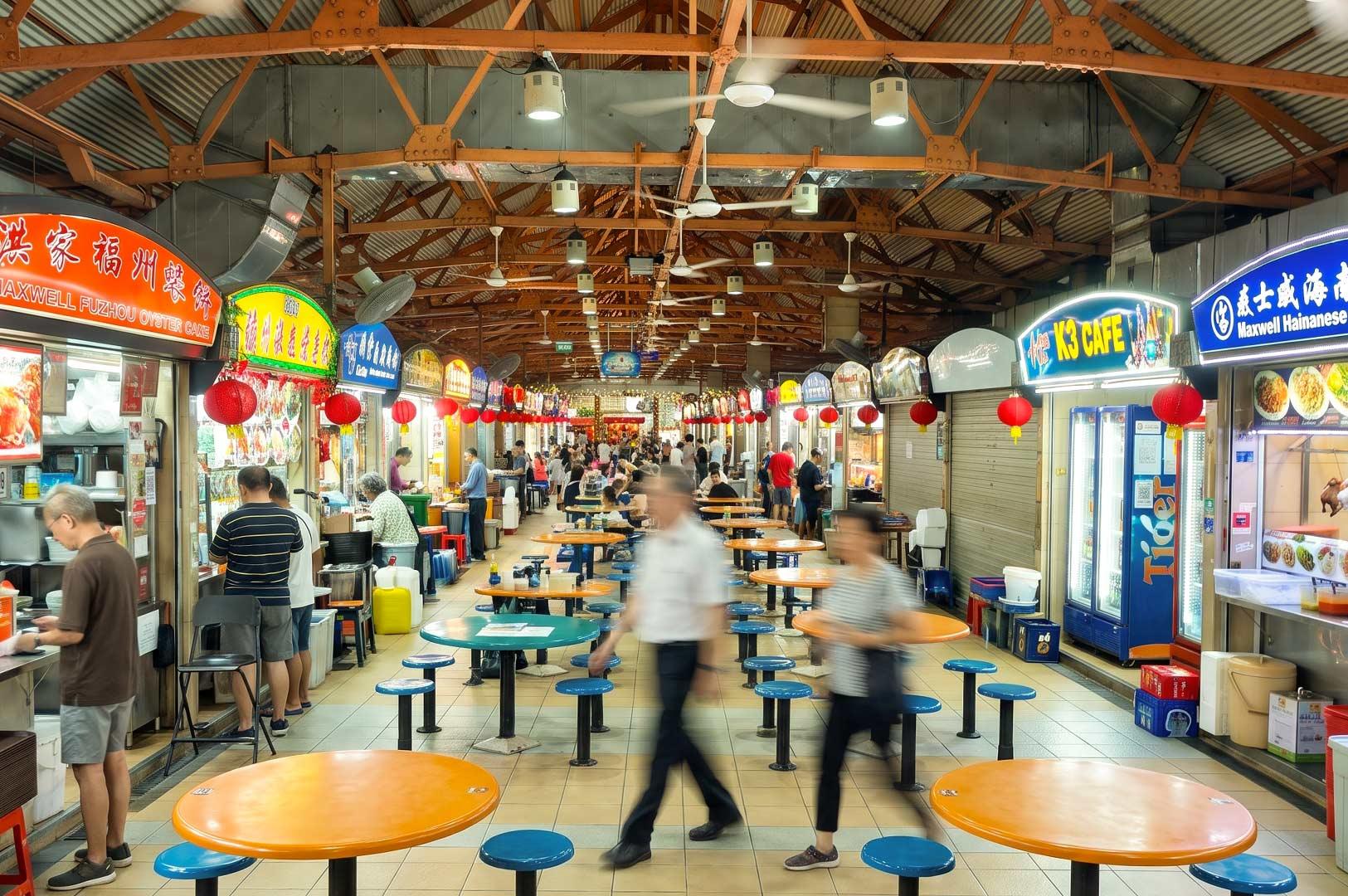 Alt text: A bustling hawker centre in Singapore with rows of colorful food stalls, round communal tables, and diners enjoying local meals. Bright illuminated stall signs, red lanterns, and an open industrial-style ceiling create a lively atmosphere as people walk through the central aisle.