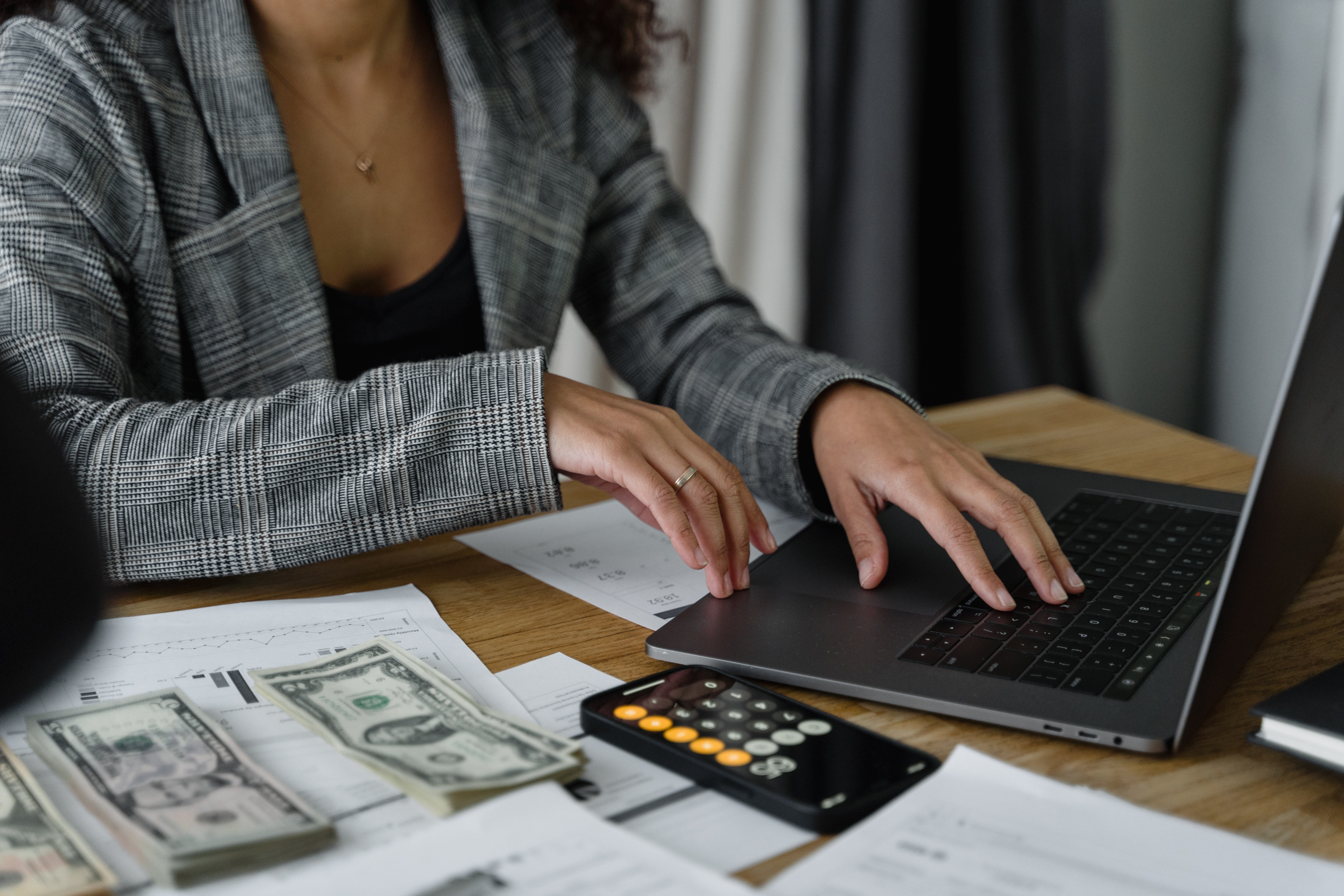 A person sitting at a table, typing into their laptop, with some cash, papers, and a calculator next to them.