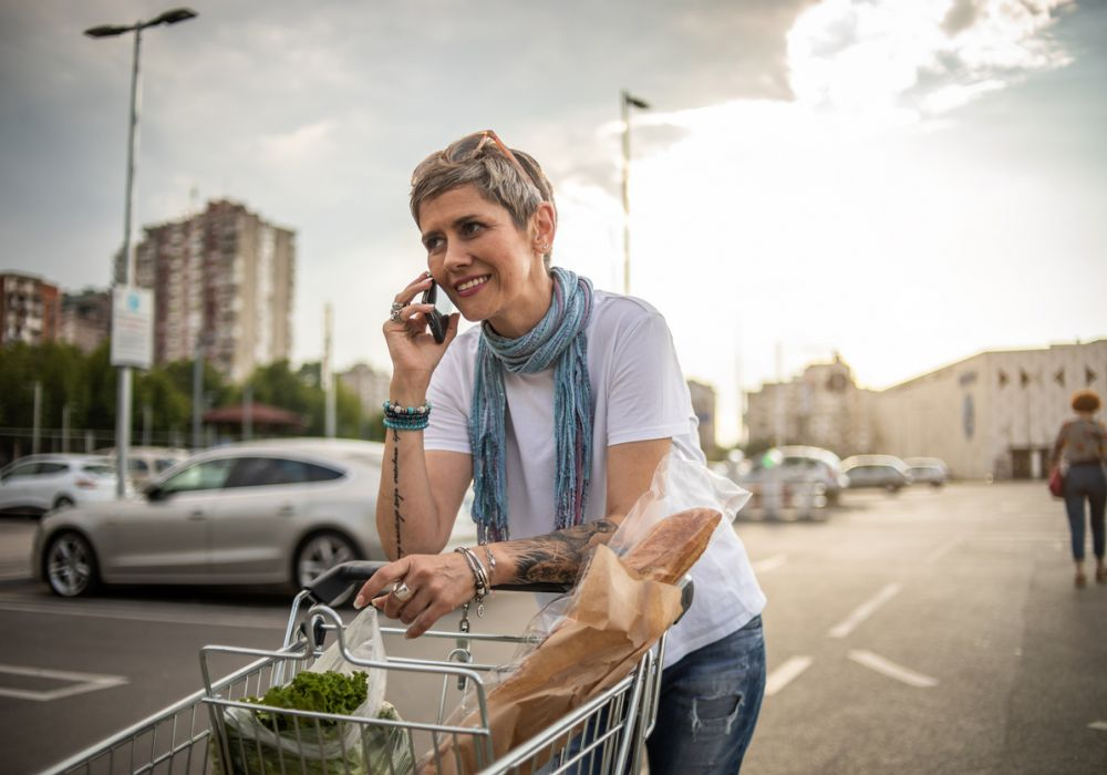 Short-haired blonde woman pushing a shopping cart through a parking lot and talking on her cell. 