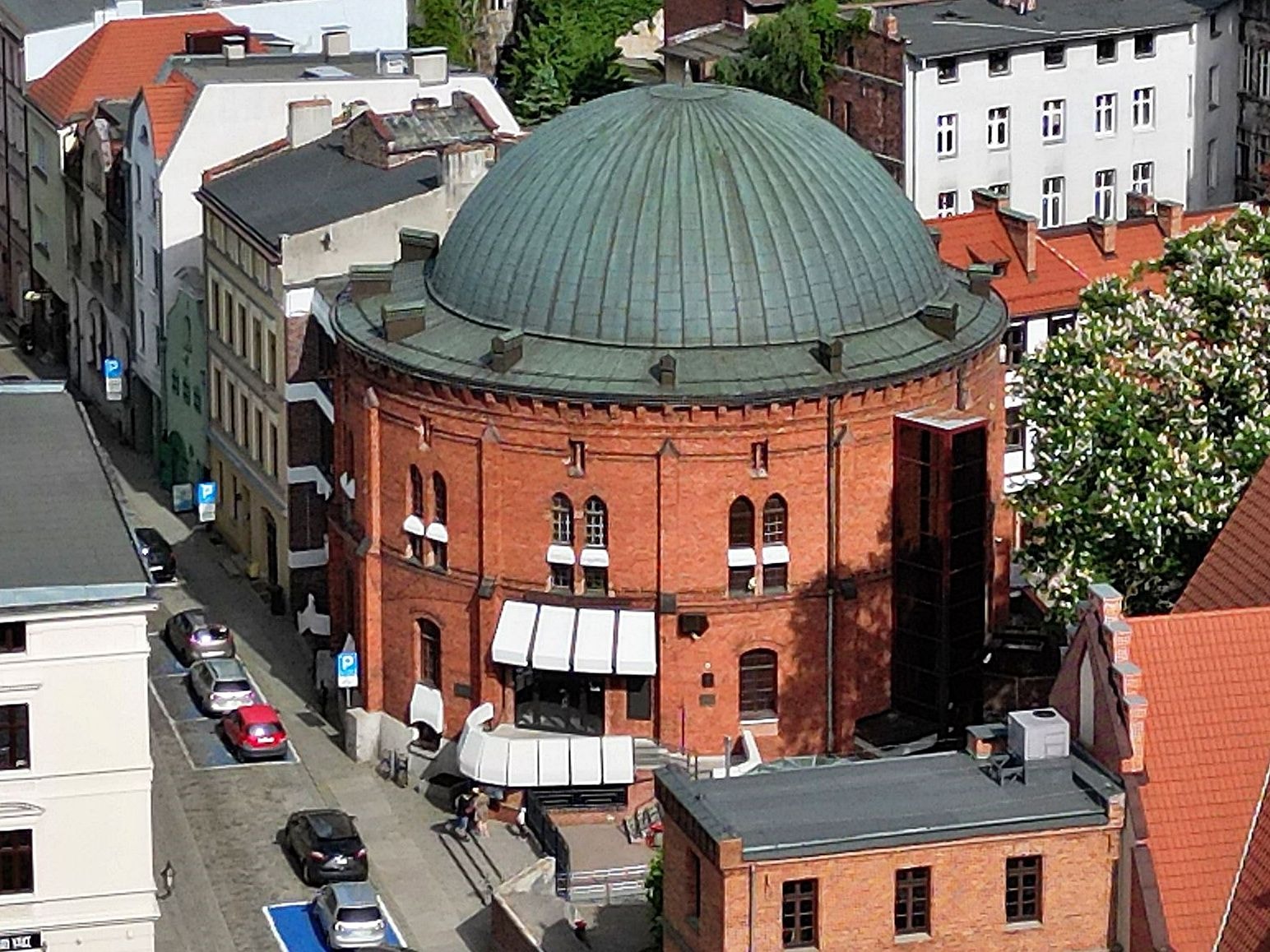 Budynek Planetarium w Toruniu. Obiekt z czerwonej cegły w kształcie rotundy otoczony wieloma budynkami starego miasta. Źródło: https://commons.wikimedia.org/wiki/File:Torun_planetarium.jpg