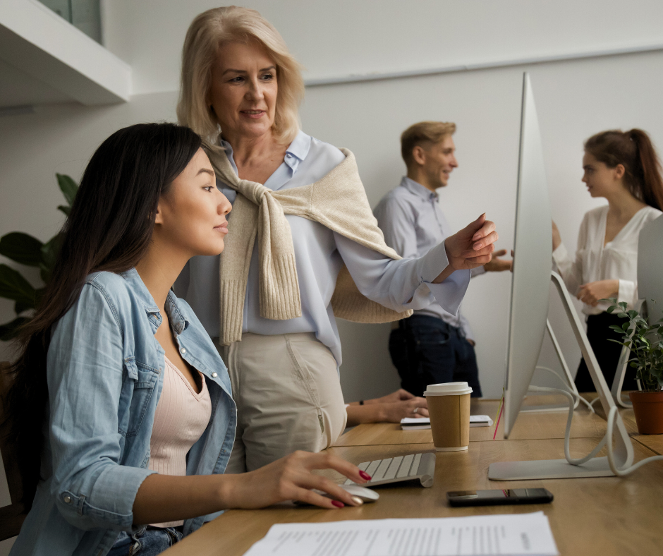 two women look at a computer in the foreground while a man and woman talk in the background