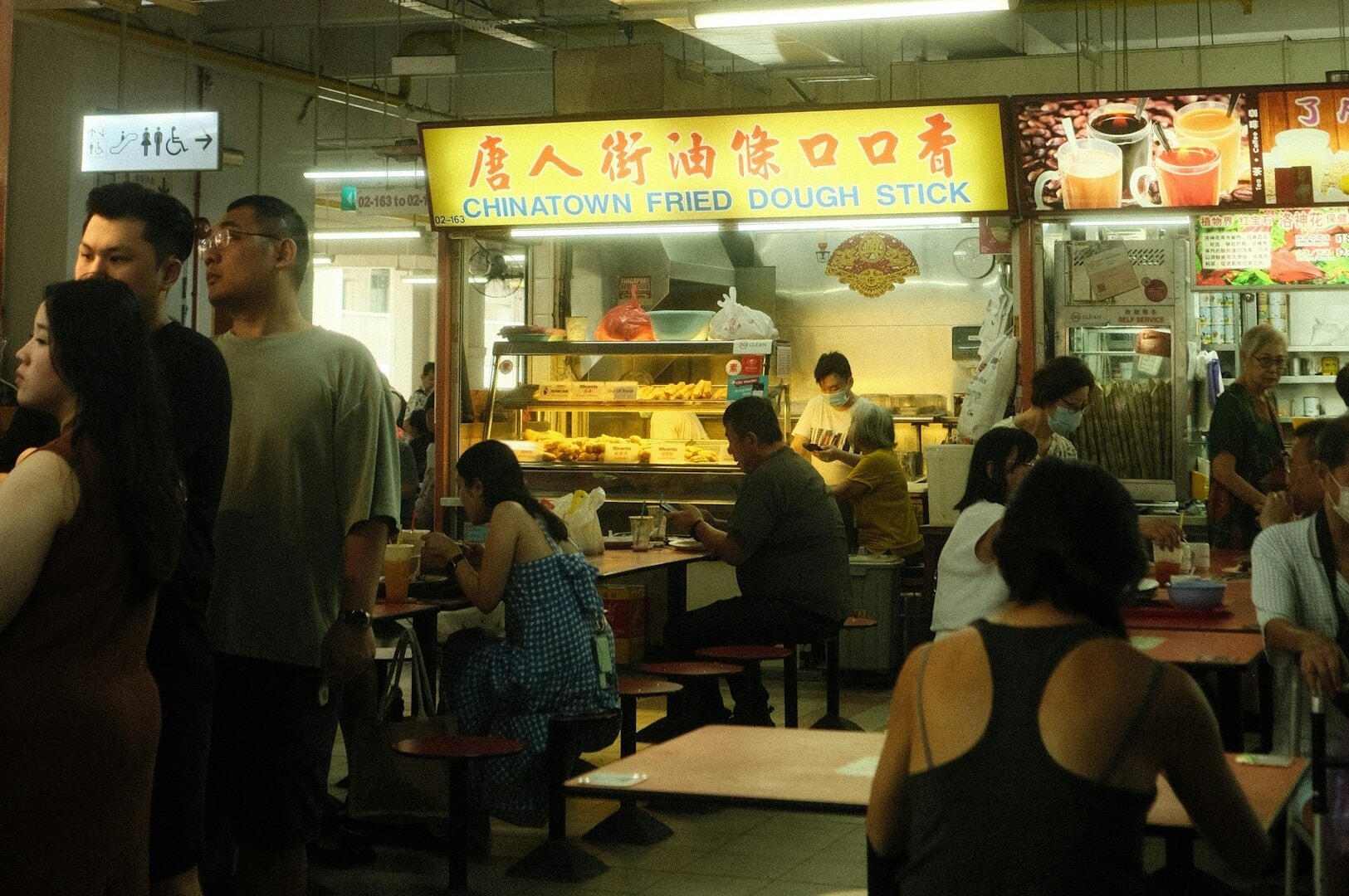A bustling food court with a "Chinatown Fried Dough Stick" stall. Diners seated at tables, engaging, and a warm, lively atmosphere.