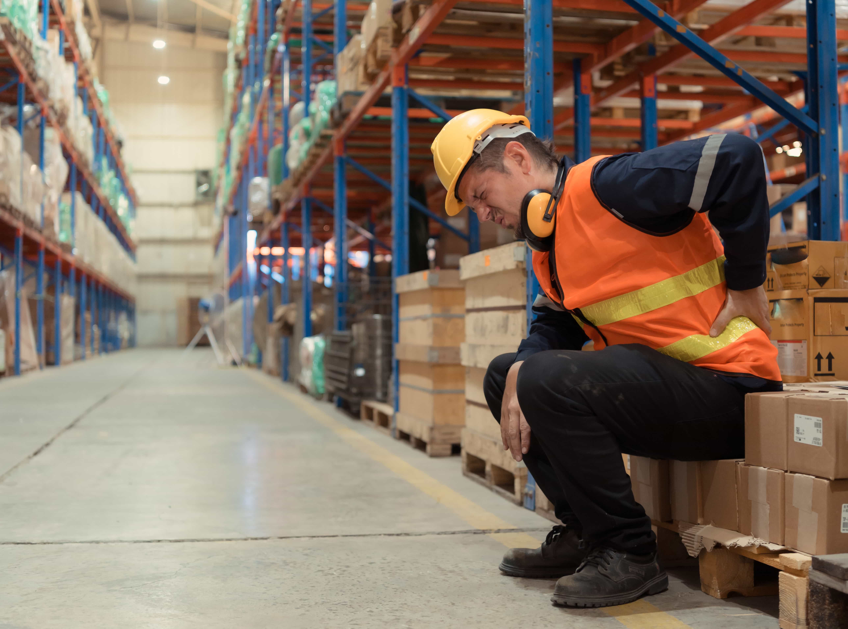 Man experiencing back pain due to repetitive strain and heavy lifting at work. He is in a warehouse.
