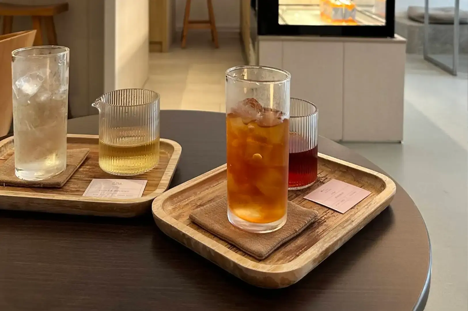 A close-up shot on a dark wooden cafe table highlights two wooden serving trays holding cold tea beverages. In the foreground, a tray holds a tall glass of iced amber tea next to a ribbed glass cup with a darker reddish liquid. To the left, a second tray presents a tall glass packed with ice alongside a ribbed glass pitcher filled with light golden-yellow tea, set against the softly blurred backdrop of the cafe counter.