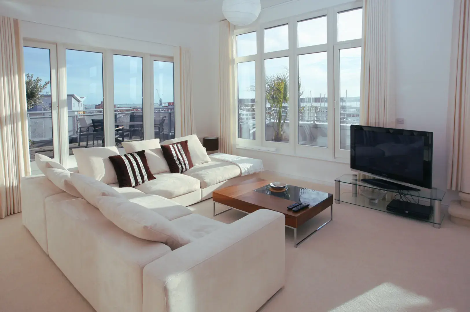 A spacious living room with a large, cream-colored sectional sofa, a wooden coffee table, and a television on a glass stand, all illuminated by natural light from large windows.