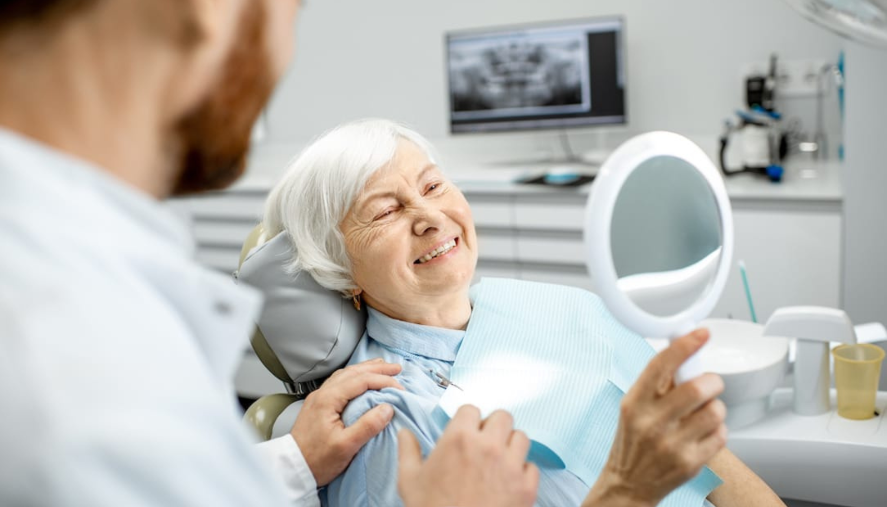 Eldre kvinne i tannlegestol smiler mens hun ser i håndspeil, med tannlege som trøster henne ved å legge hånden på skulderen.