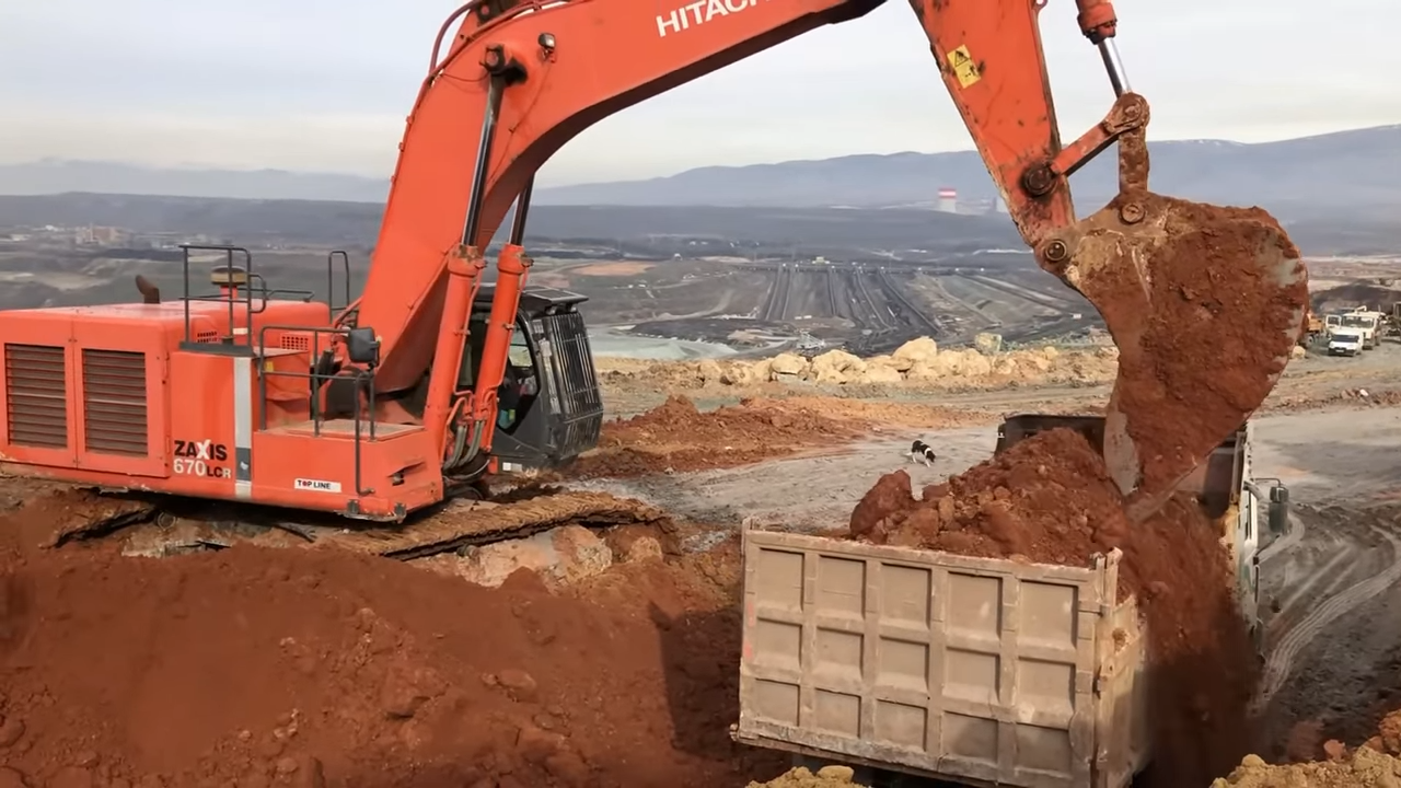An orange Hitachi ZAXIS 670 LCR hydraulic excavator in the process of loading dirt into the back of a large industrial dump truck at a mining site. The excavator's bucket is caught mid-motion, spilling soil. The bac