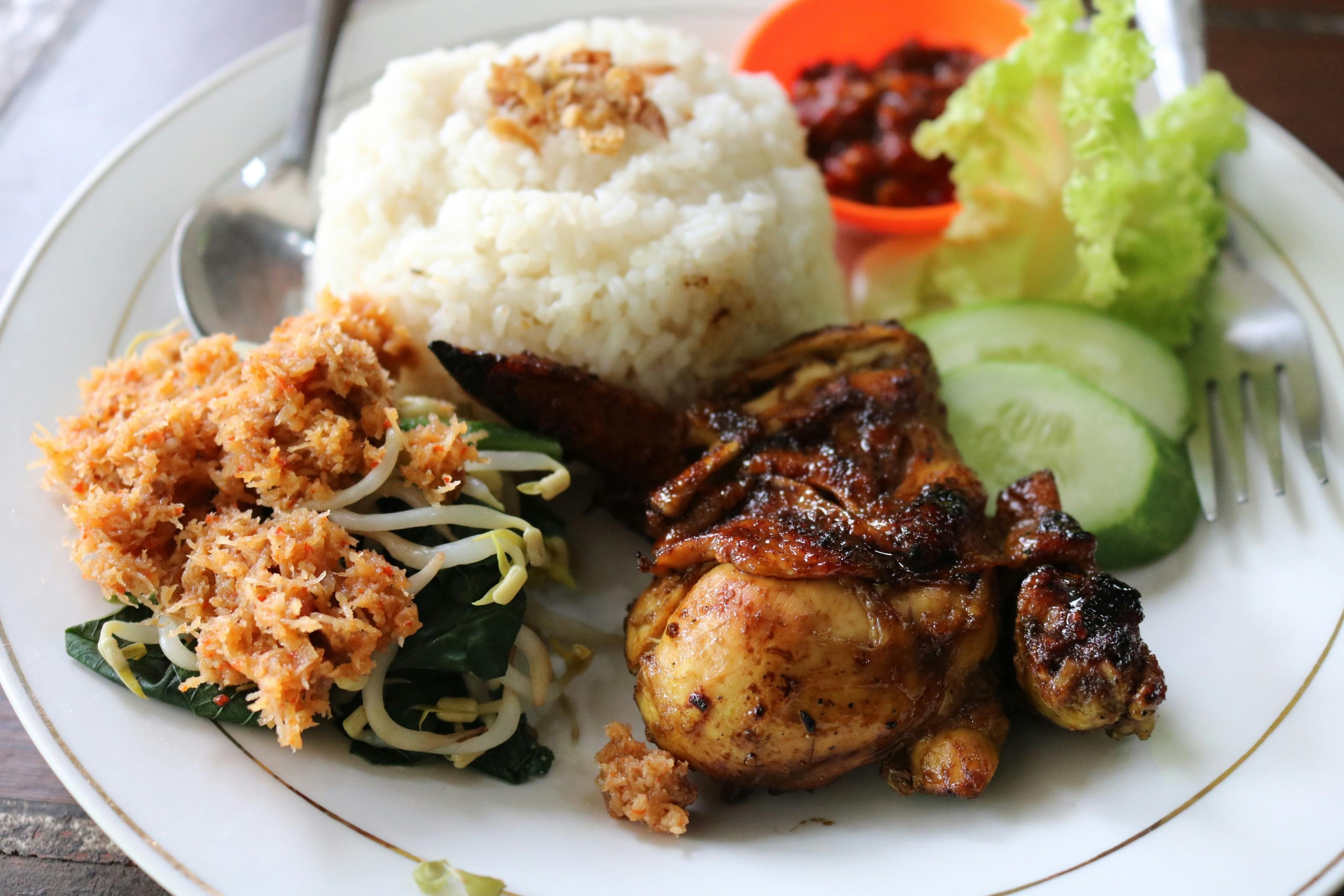 This plate features a hearty serving of Indonesian grilled chicken (Ayam Bakar) served alongside a mound of white rice topped with fried shallots. The meal is complemented by a side of urap (vegetable salad with spiced coconut), fresh cucumber slices, lettuce, and a small dish of spicy sambal.