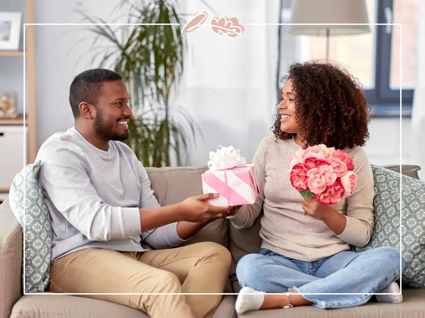 A happy couple sitting on a couch exchanging gifts, the woman holding a pink flower bouquet and the man presenting a pink wrapped gift box. Fabulous Flowers and Gifts.