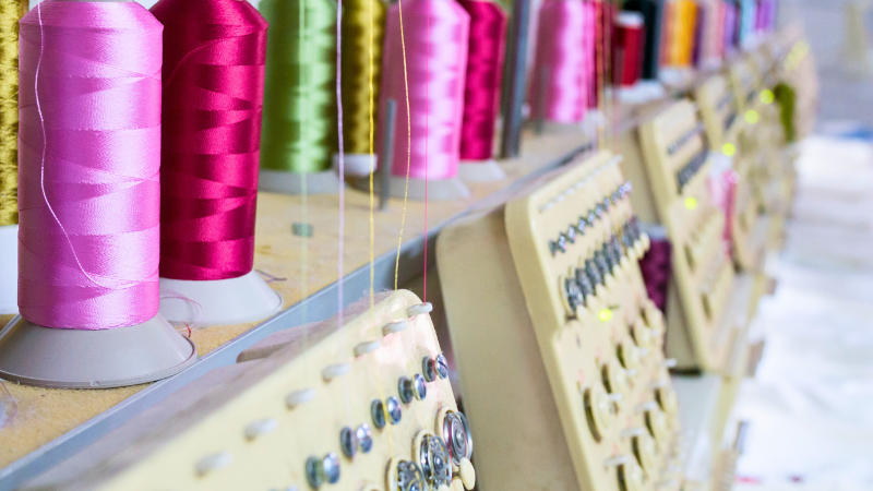 A row of spools of colorful polyester thread, pink, magenta, and green, are mounted on an embroidery machine.