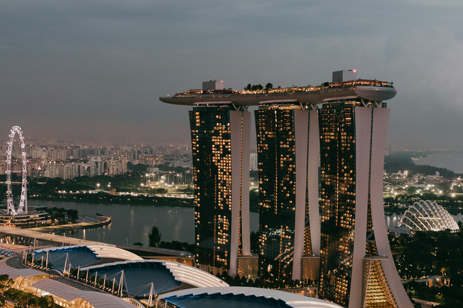 Aerial view of Marina Bay Sands in Singapore at dusk, with illuminated windows and rooftop gardens. The Singapore Flyer is visible on the left.