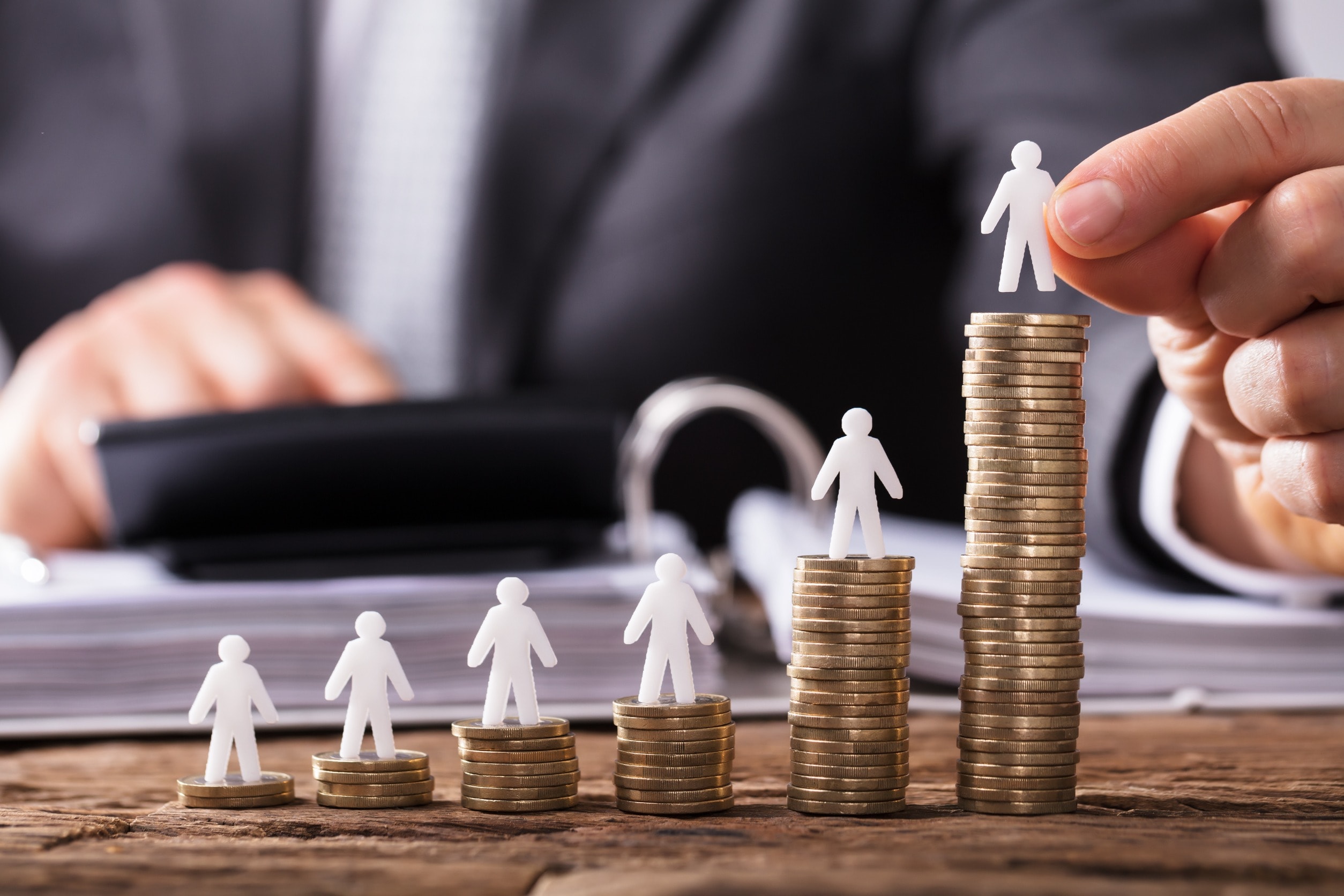 Six piles of coins sit on a desk with small paper figurines on top