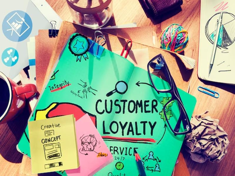 A green pad with "Customer Loyalty" written on it, surrounded by eyeglasses, paper clips, a coffee mug, and other office items, creating a workspace scene focused on business strategy.
