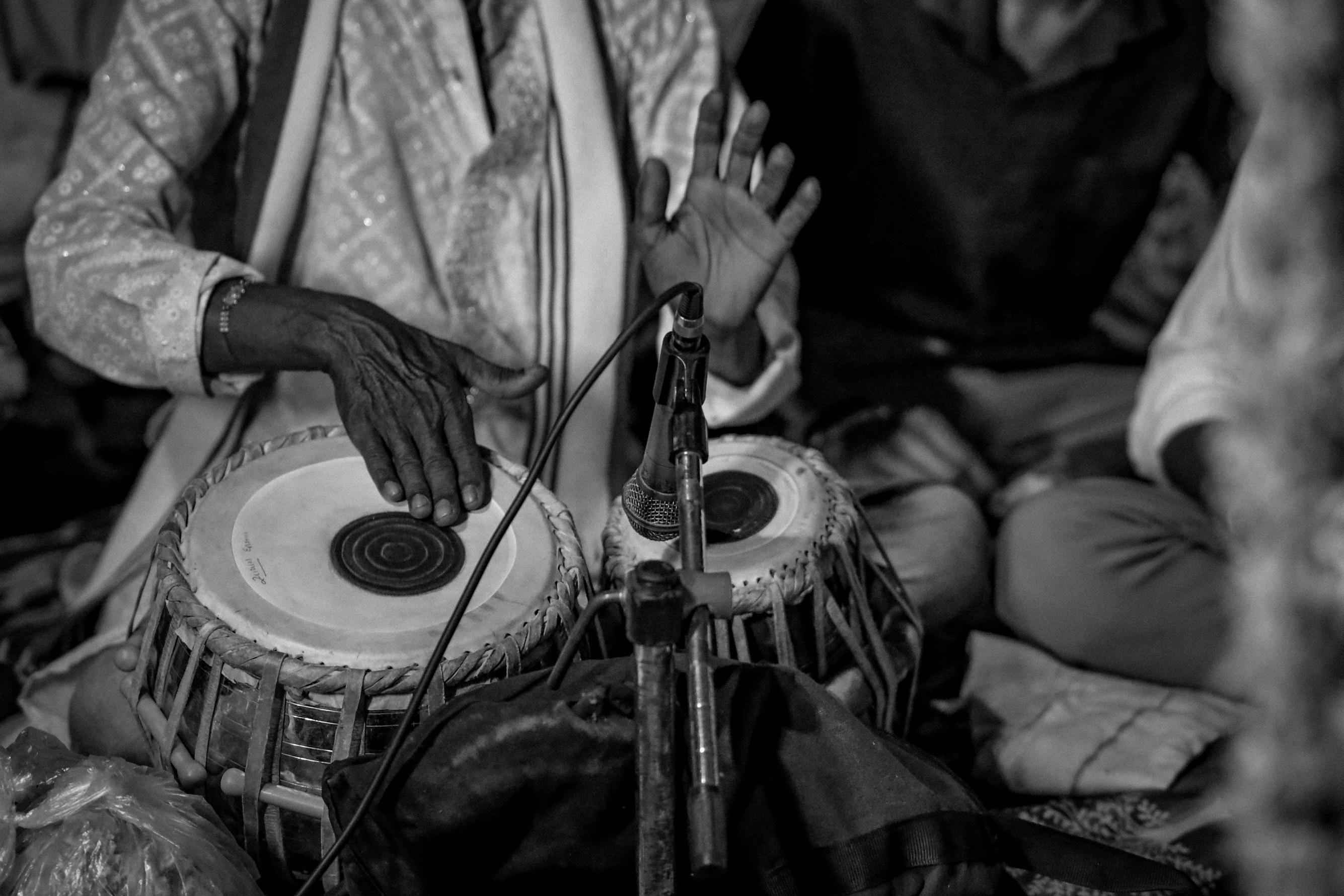 A close-up black-and-white shot highlights the skilled hands of a musician playing the tabla, with fingers poised to strike the drumheads. A microphone is positioned directly in front of the instrument to capture the performance, while the musician is dressed in a textured traditional outfit.