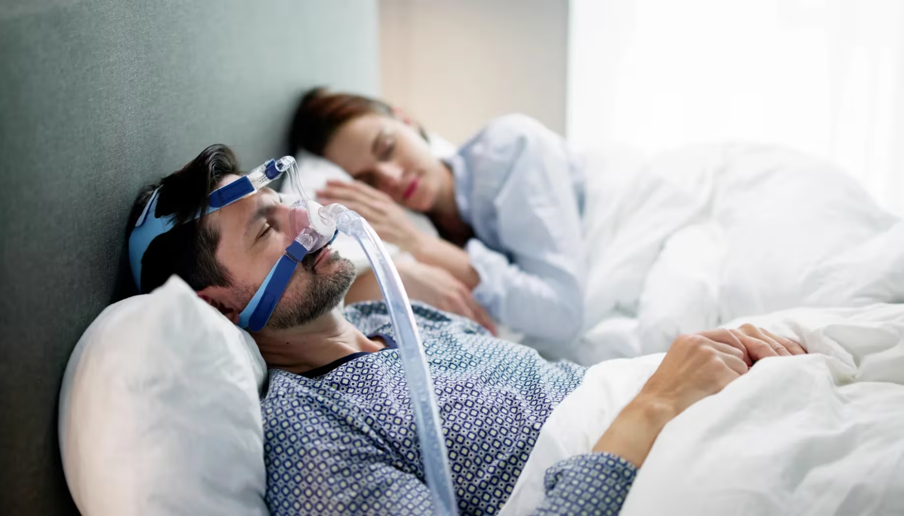 Mann med CPAP ved siden av partner; begge sover under hvitt sengetøy.