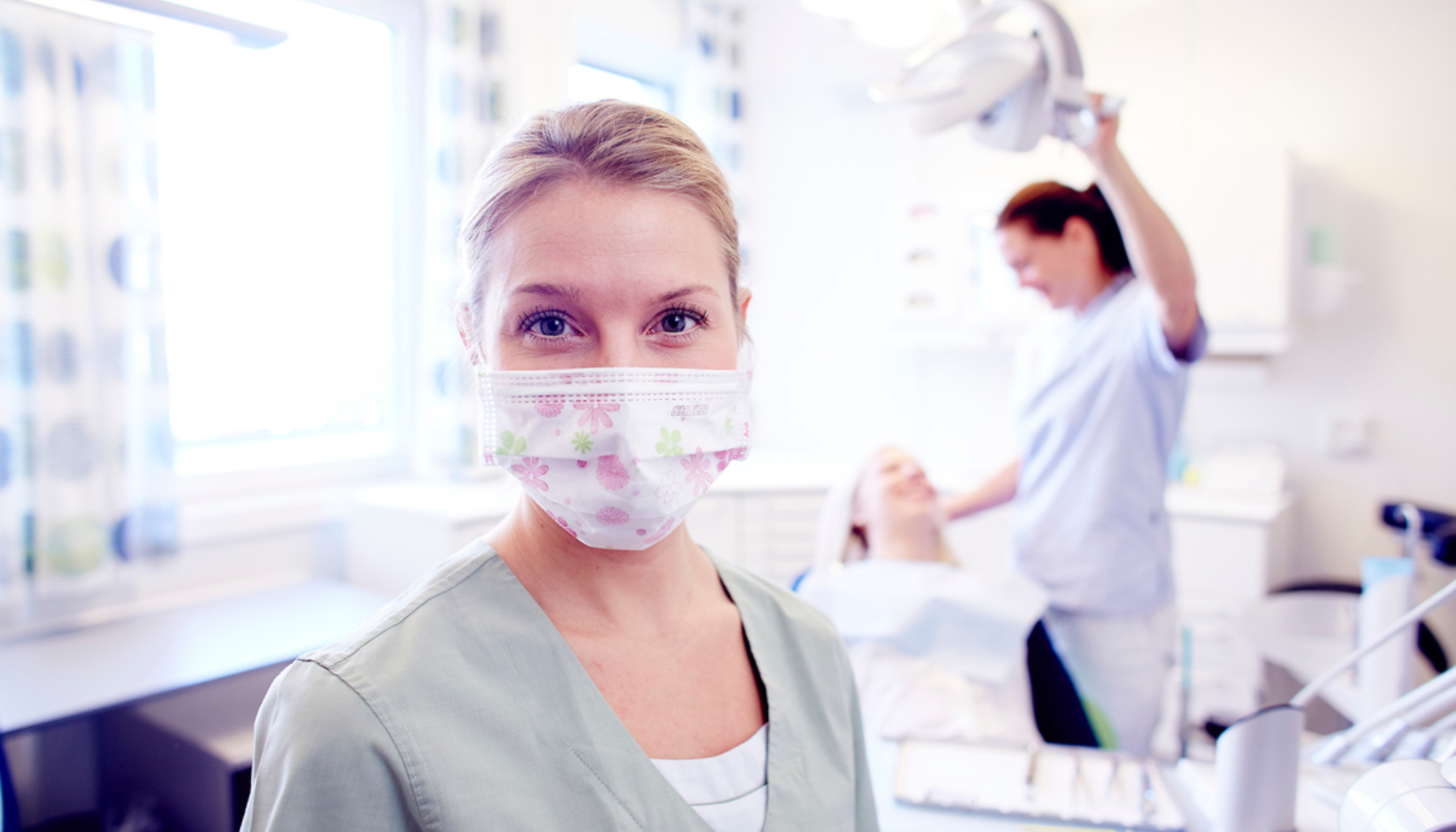 "Tannlege med munnbind smiler mot kamera, mens en pasient behandles i bakgrunnen i en lys og moderne tannklinikk."