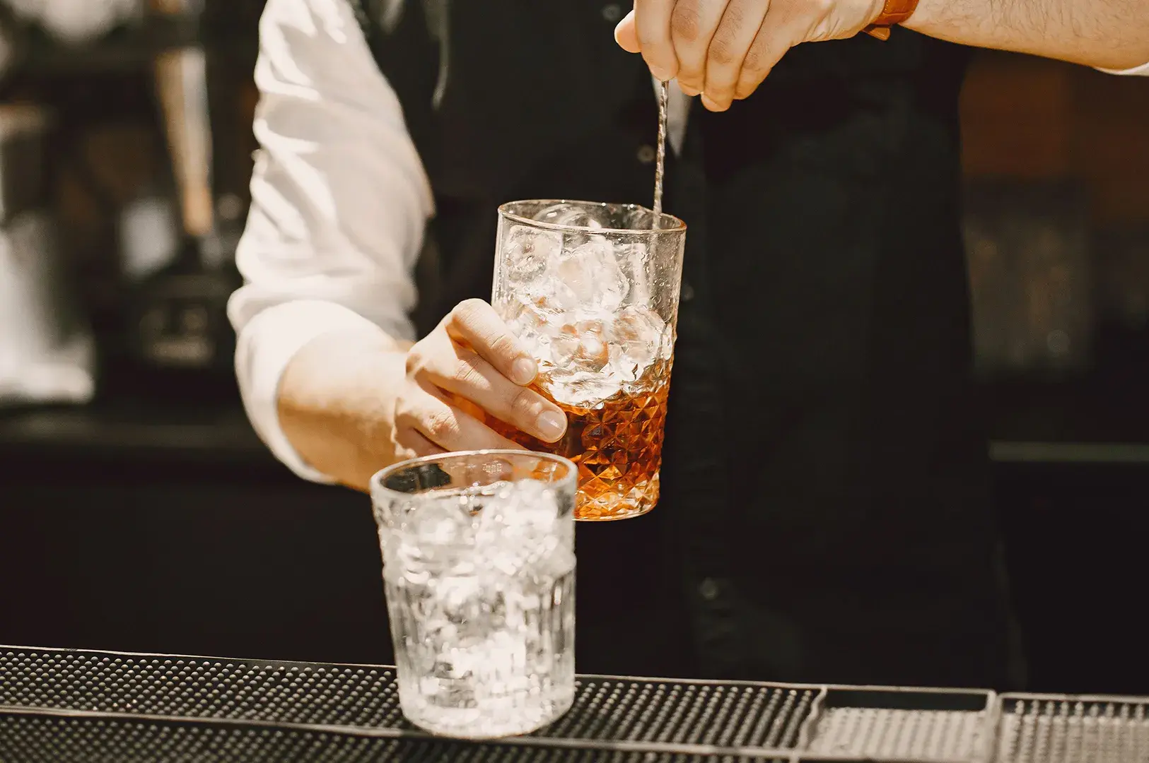 Bartender stirring an iced cocktail in a textured glass with a long bar spoon behind a bar counter.