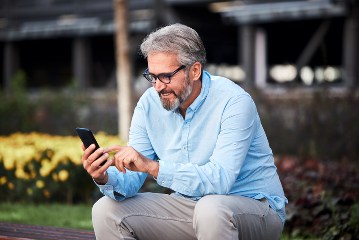 Man with gray hair and black glasses siting outside sending a text. 