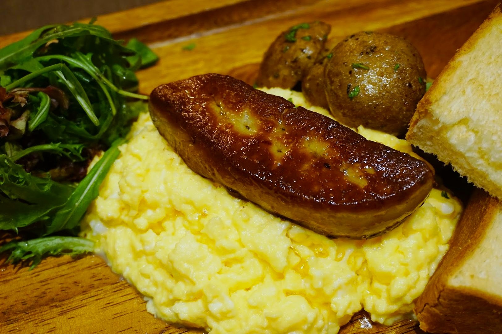 A wooden plate featuring creamy scrambled eggs topped with seared foie gras, roasted potatoes, fresh mixed greens, and slices of toasted bread.