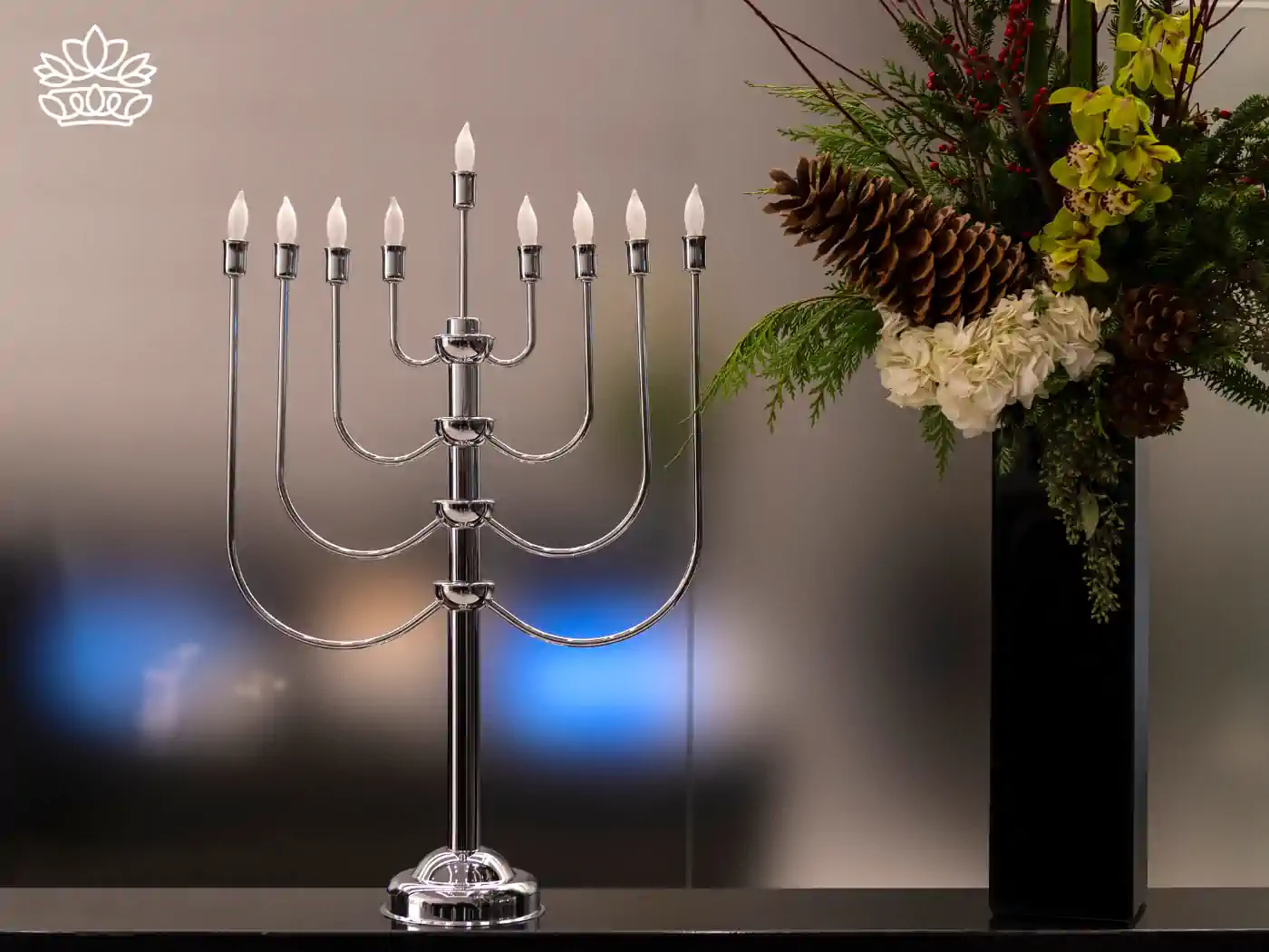 A modern silver menorah with all nine candles lit, placed next to a vase with a festive floral arrangement including pinecones and green foliage. Fabulous Flowers and Gifts - Hanukkah Flowers.