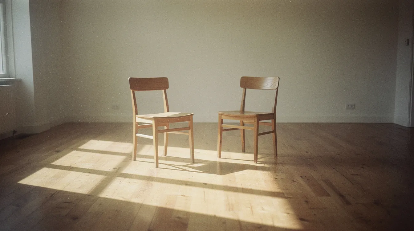 In a sunlit, empty room, two simple wooden chairs are angled toward each other, casting long, soft shadows on the warm wooden floor. The minimalist composition evokes a calm and intentional mood, reminiscent of the therapeutic space where one might explore core emotional needs and coping styles in schema therapy.