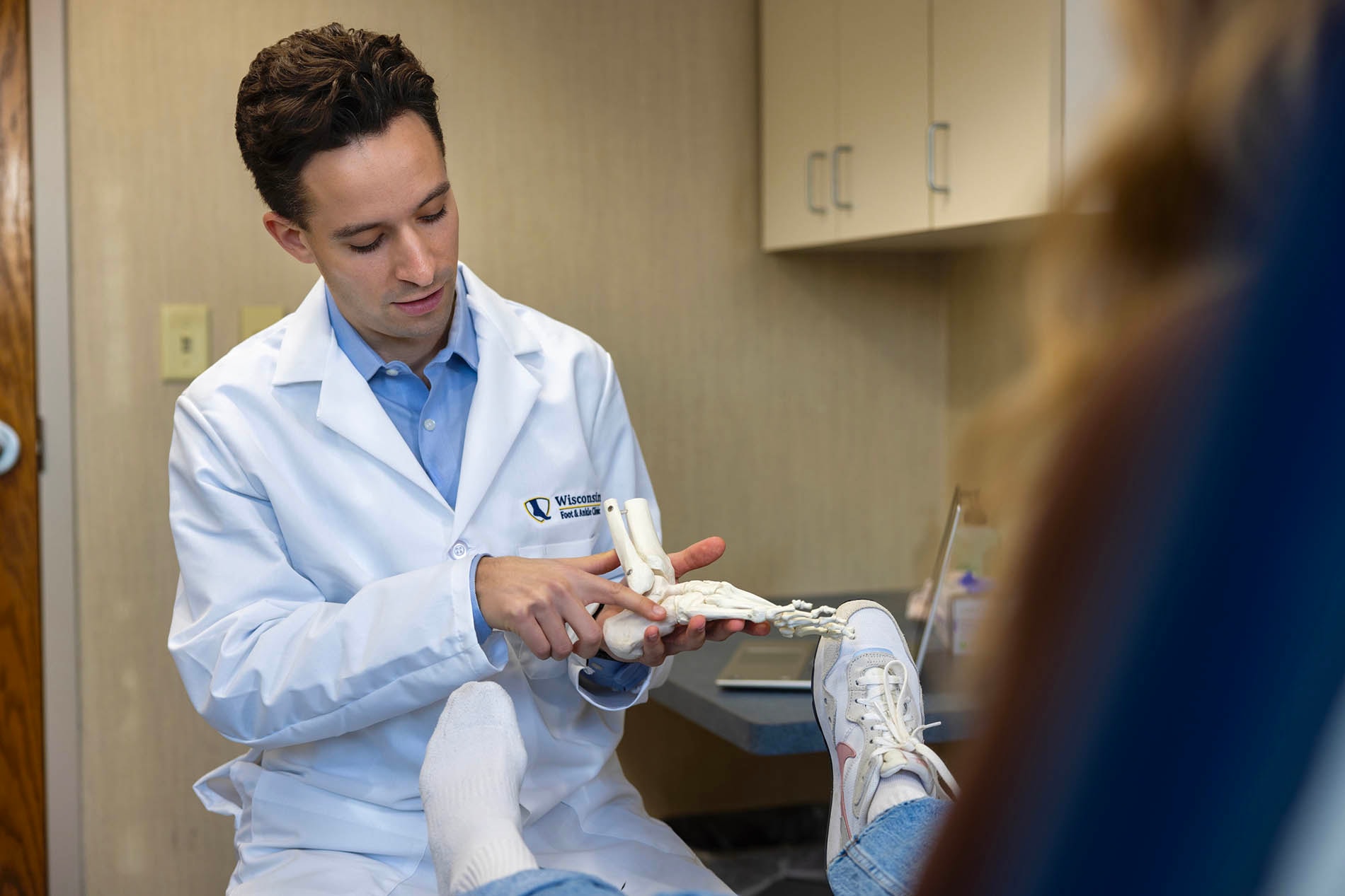 Dr. McDonald, a foot and ankle doctor at Wisconsin Foot & Ankle Clinic, sits in an exam room and uses a plastic foot and ankle bone model to explain diagnosis and treatment options to a patient, while the patient rests one leg wearing a sneaker and has the other foot in a white sock, showing a calm in-office consultation focused on understanding the problem and next steps.