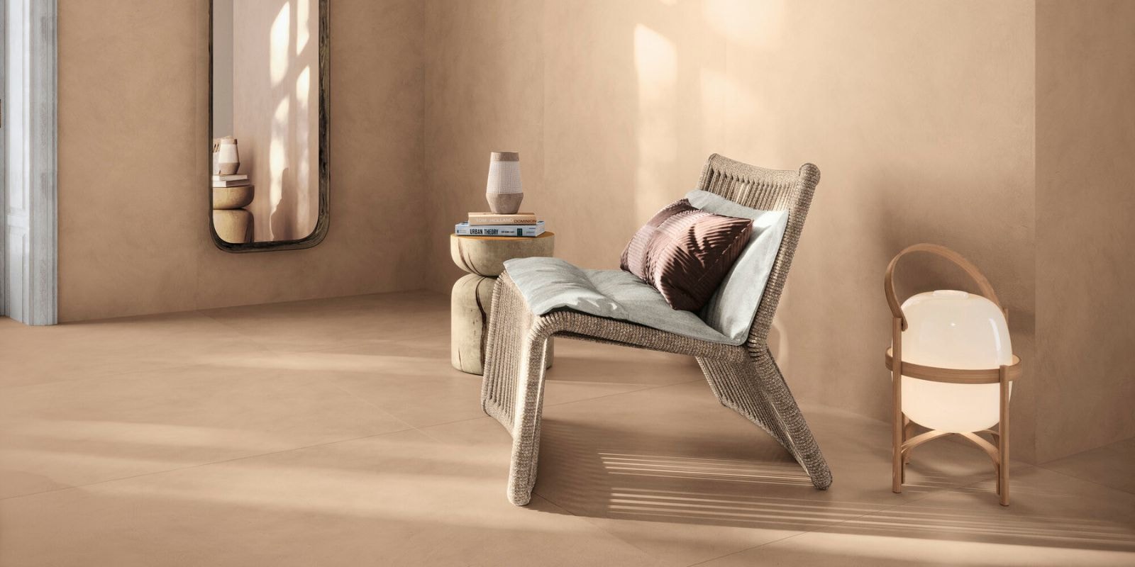Minimalist reading nook with a woven lounge chair, sculptural side table, modern lantern lamp, and a tall mirror on beige-toned walls and flooring.