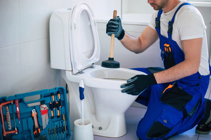 Handwerker in blauer Arbeitskleidung verwendet einen Pümpel an einem Toilettenbecken.