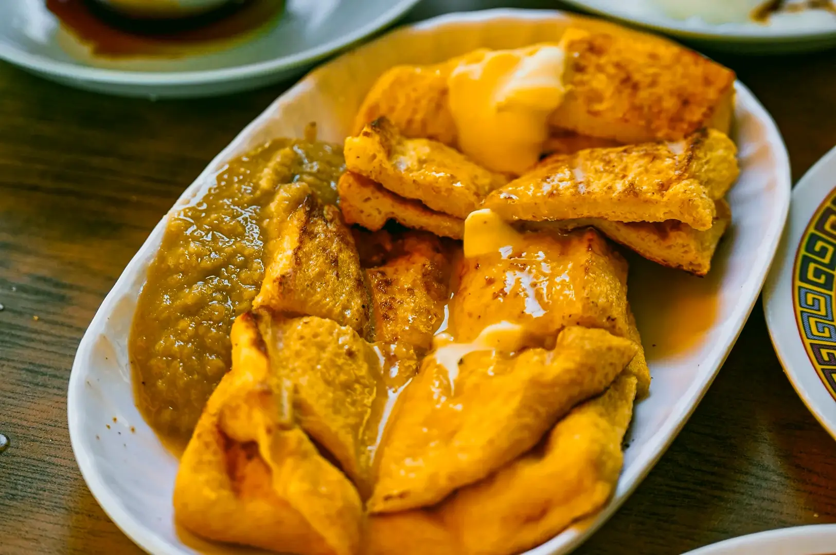 Close‑up overhead shot of a traditional Singapore breakfast dish featuring a folded omelette topped with thick toast pieces and golden sauce, served on an oval plate alongside a thick brown condiment on a wooden table.