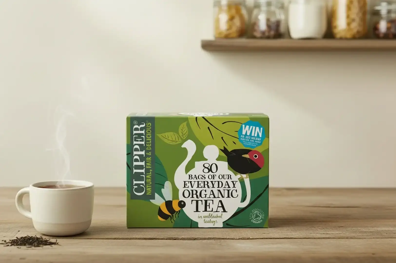 A warm, sunny kitchen setup centered around a green box of Clipper "80 Bags of Our Everyday Organic Tea," which features whimsical illustrations of a teapot, a bumblebee, and a red-headed bird. A small pile of loose tea leaves rests to the left of a steaming white coffee mug on a rustic wooden countertop, with glass pantry jars visible on a floating shelf overhead.