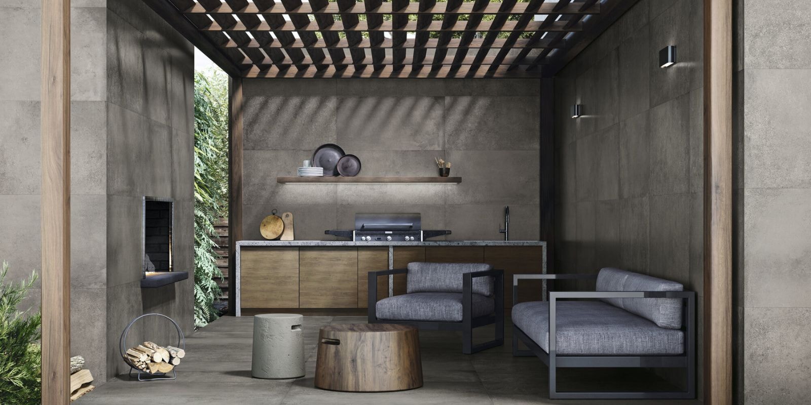 Modern outdoor kitchen and lounge area with large-format grey concrete tiles on the floor and walls, wood cabinetry, a sleek built-in grill, minimalist floating shelf, and lounge furniture with black metal frames and grey cushions, all under a wooden pergola.