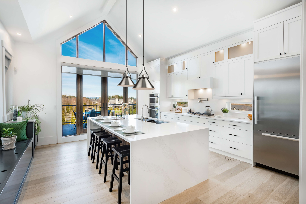 Pendant light fixtures with an industrial look hang above a white kitchen island in a bright modern kitchen with black stools and tall windows.