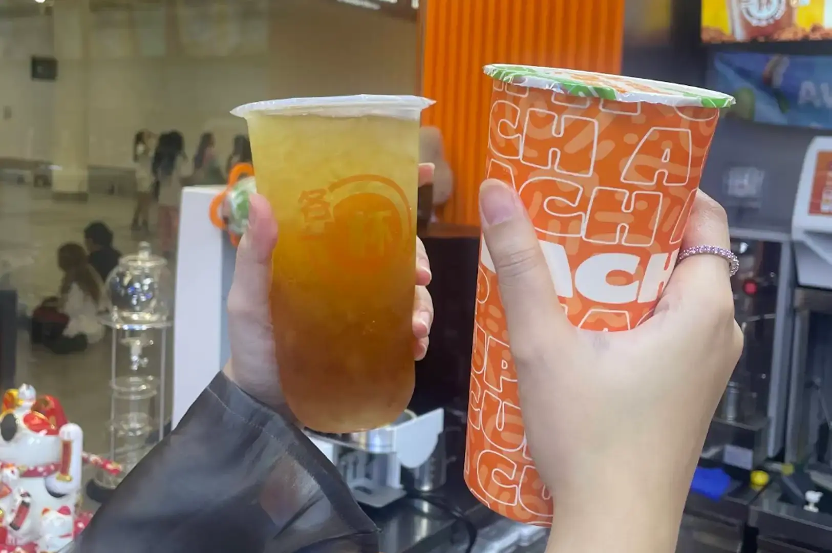 Two hands hold up bubble tea cups in a celebratory toast, with one person holding a clear plastic cup filled with iced golden fruit tea showcasing the Each A Cup logo, and the other holding a tall paper cup decorated with a vibrant orange graphic typography pattern.